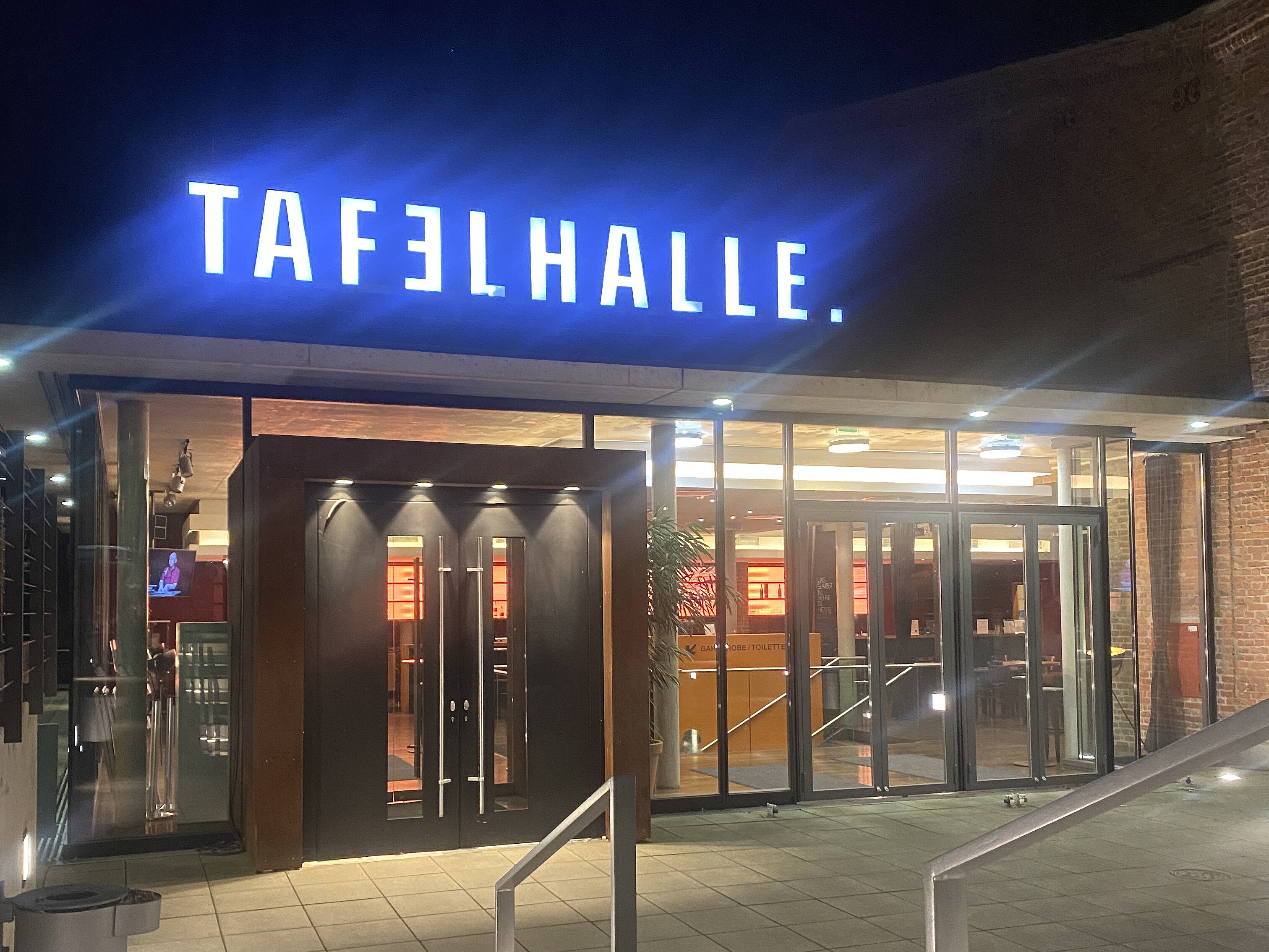 Eingang der Tafelhalle bei Nacht, mit blau leuchtendem Neon-Schriftzug über der Glastür.