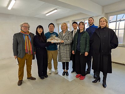 Gruppe von acht Personen in einem leeren Atelierraum, eine Person hält ein mit Folie verpacktes Brot, helle Wände und Fenster im Hintergrund.