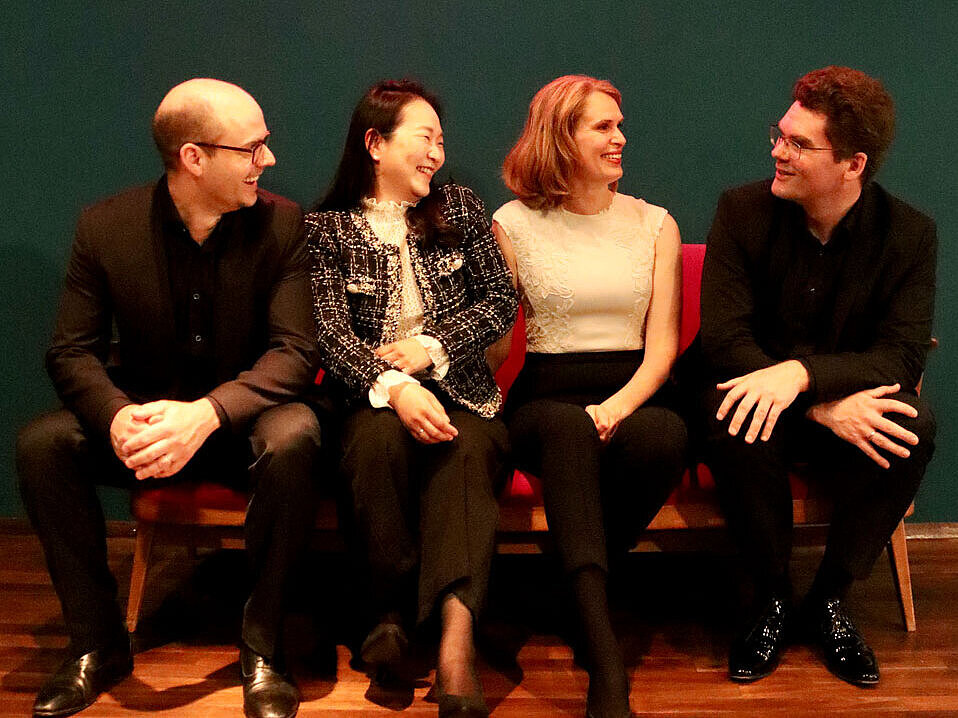 zwei Männer und zwei Frauen sitzen auf einer Couch vor einer dunkelgrünen Wand, darüber die neonfarbene Schrift Tafelhalle