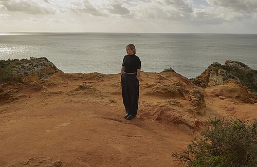 Frau in schwarz gekleidet auf felsigem Berg mit dem Meer im Hintergrund