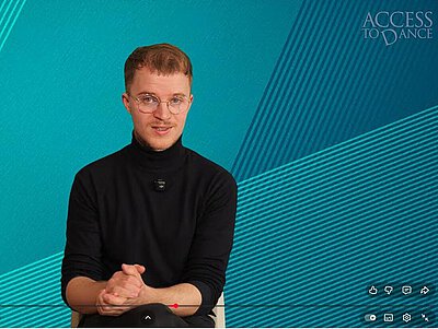 Person mit kurzem Haar sitzt vor blau-grünem Hintergrund und trägt schwarzen Pullover mit Mikrofon am Kragen.