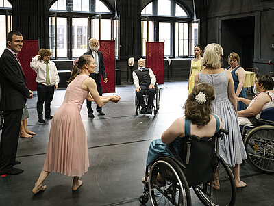 Gruppe von Menschen, darunter mehrere im Rollstuhl, in einem großen Raum mit hohen Fenstern, einige tragen festliche Kleidung und eine Frau in einem rosa Kleid macht eine einladende Geste.
