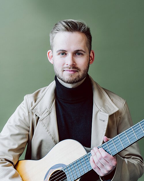 junger Mann in schwarzem Rollkragenpulli und beigem Mantel mit Gitarre