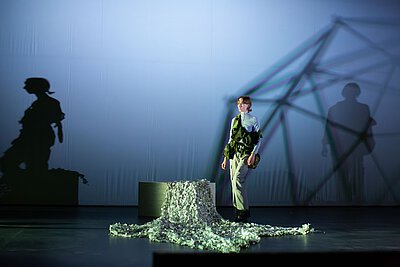 Person mit einer militärischen Weste, die über die Bühne geht. Im Hintergrund eine weiße Leinwand, auf die Schatten der Person und einer geometrischen Form geworfen werden. Die Beleuchtung ist hell und diffus, mit einem bläulichen Farbton. Die Person schaut fokussiert in eine Richtung. Die Atmosphäre ist düster und ernst.