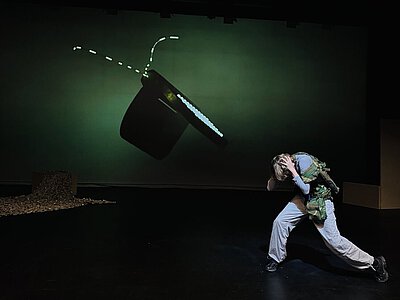 Person auf dunkler Bühne in dynamischer Pose vor grünem Hintergrund mit projizierter Animation eines leuchtenden, schwebenden Objekts.