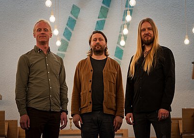 Drei Personen stehen nebeneinander in einem Raum mit hängenden Glühbirnen und blauen Wanddekorationen im Hintergrund.