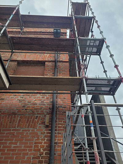 Blick nach oben auf ein mehrstöckiges Gerüst vor einer Backsteinwand mit Metallrohren und Holzbohlen