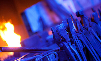 Das Foto zeigt ein brennedes Feuer in der Schmiede-Esse und davor hängend eine Reihe an Schmiedewerkzeug