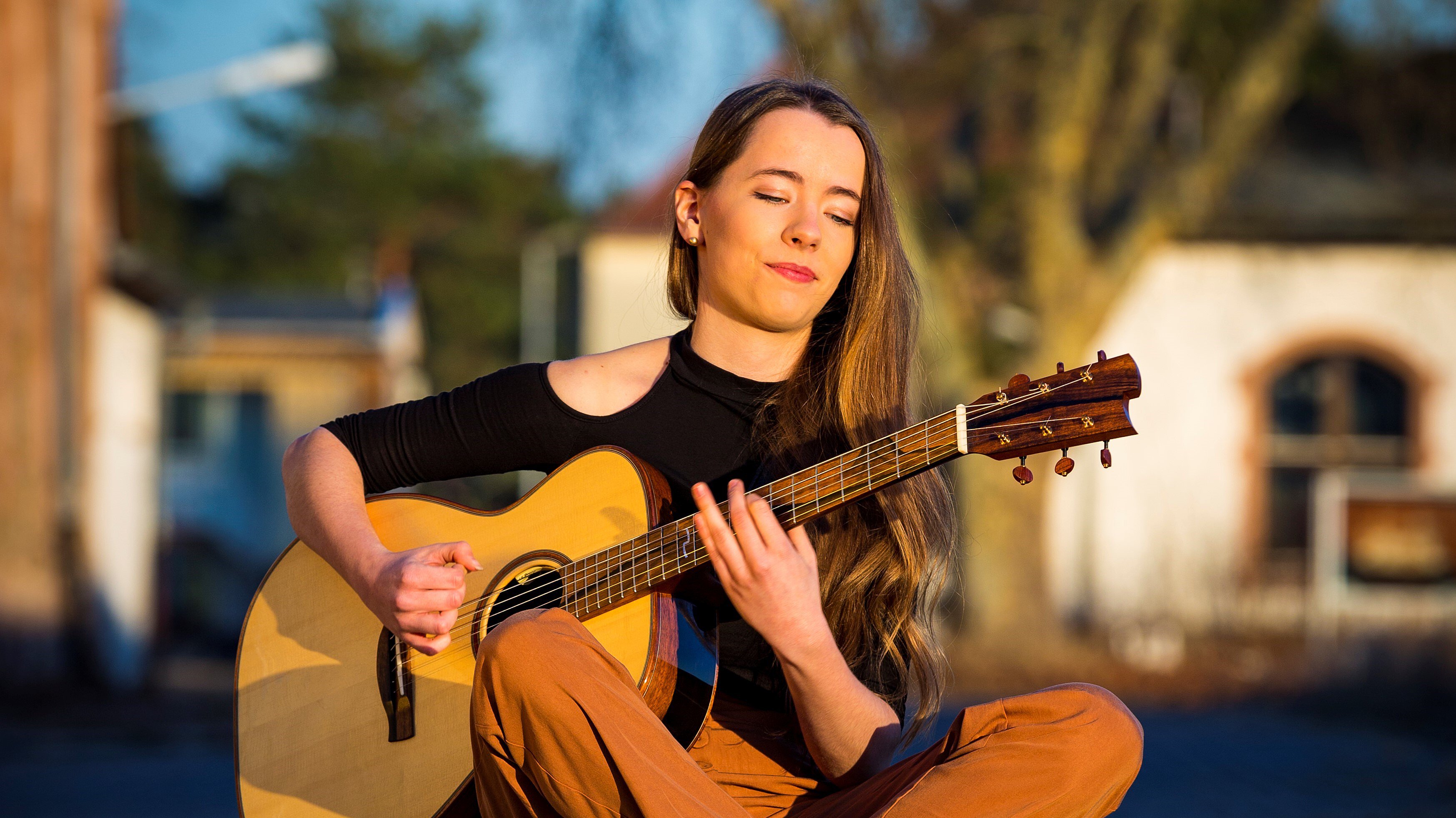 Musikerin sitzt im Schneidersitz draußen mit geschlossenen Augen und spielt akustische Gitarre